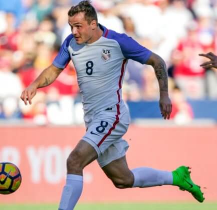 An American soccer player runs forward with the ball as an opposing defender follows him.