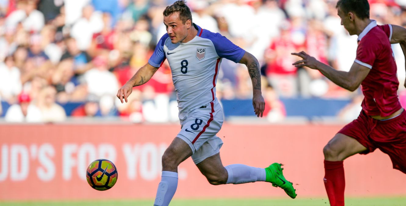 An American soccer player runs forward with the ball as an opposing defender follows him.