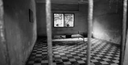 black and white picture of a jail cell with a cot and bare, concrete walls behind bars.